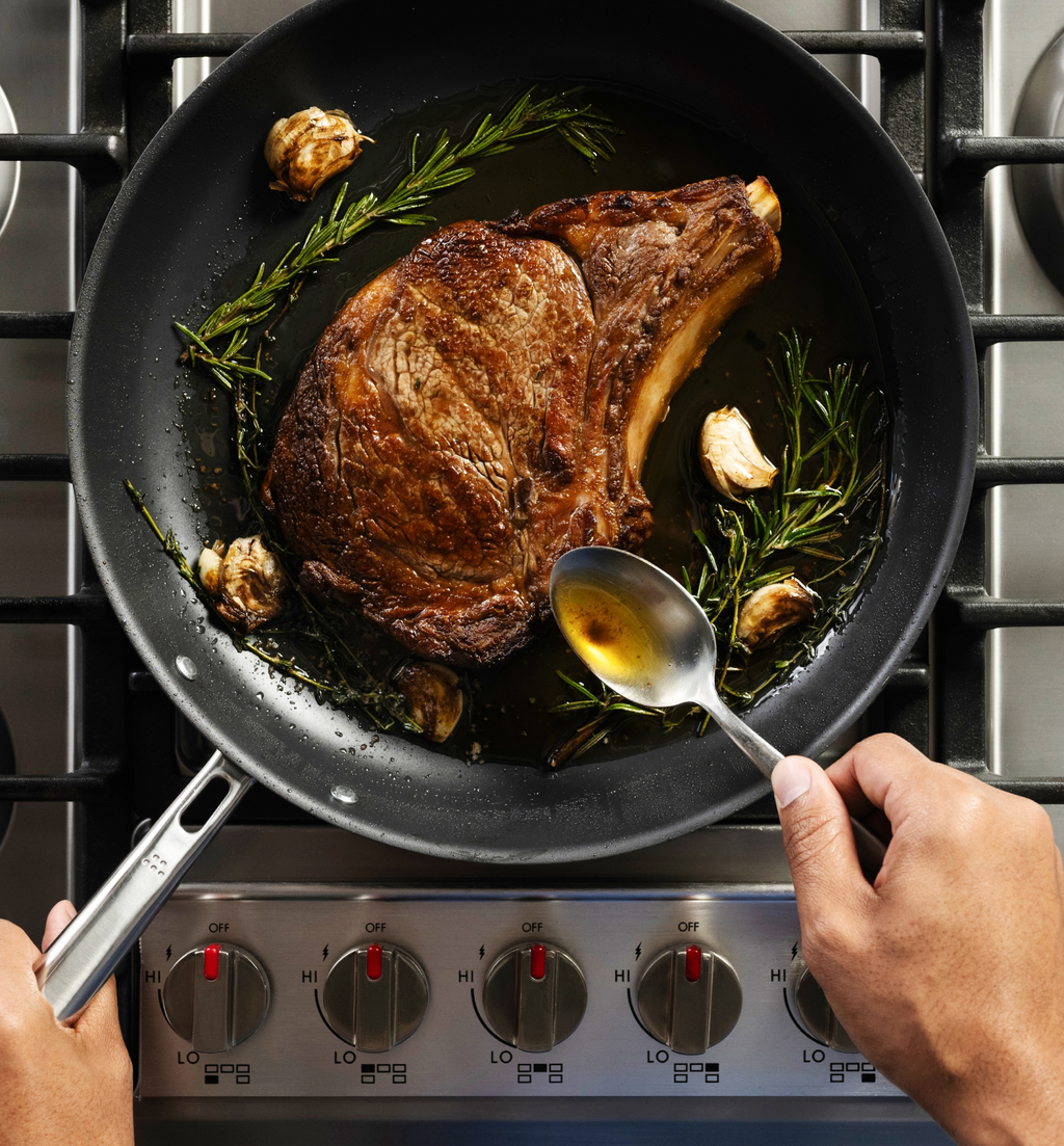 Basting a bone-in ribeye steak with butter, rosemary, and garlic in a skillet